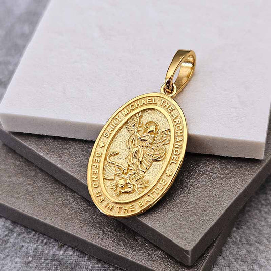 A gold Saint Michael the Archangel pendant with an embossed design, resting on a gray textured surface with a white marble block behind it.
