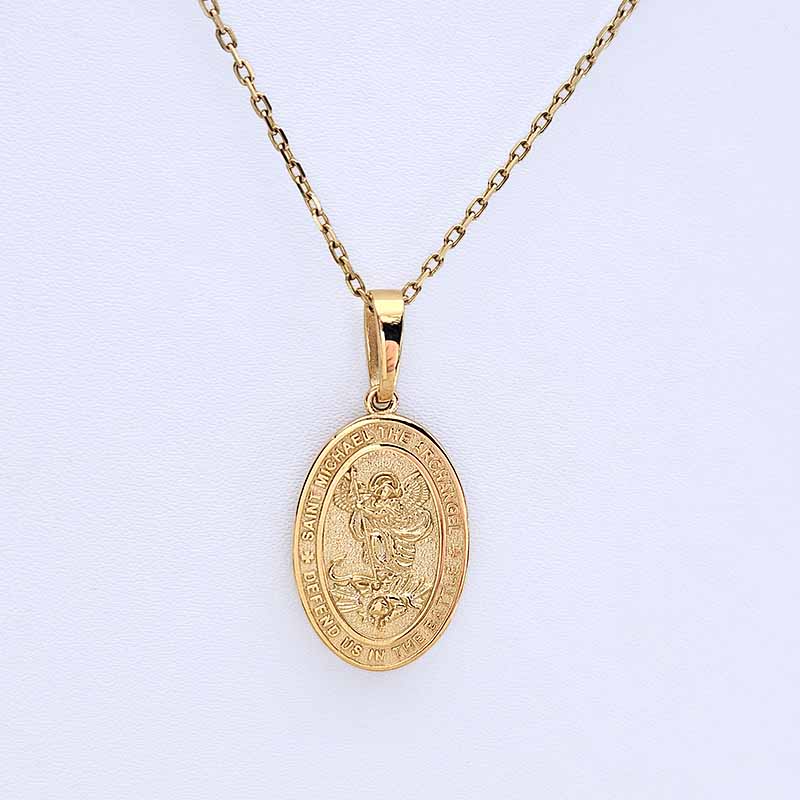 A gold oval pendant necklace featuring an embossed religious design of St Michael, suspended from a delicate gold chain against a light gray background.