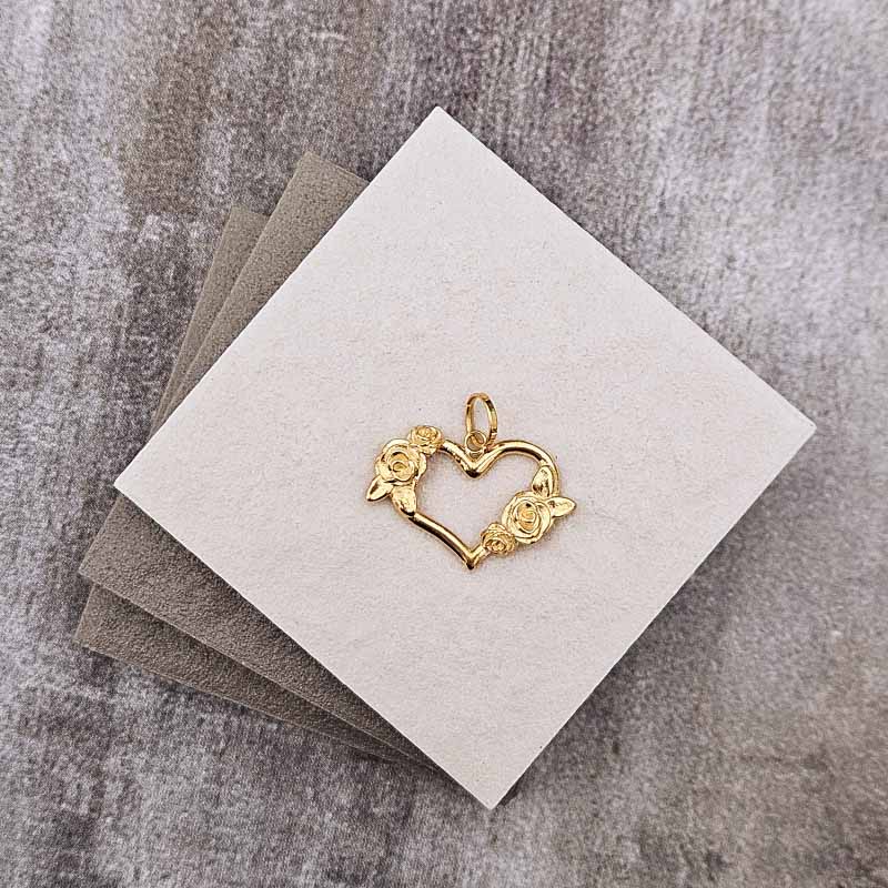 A small gold heart-shaped pendant with two rose designs, displayed on a square cream card over stacked gray cards on a textured gray surface.