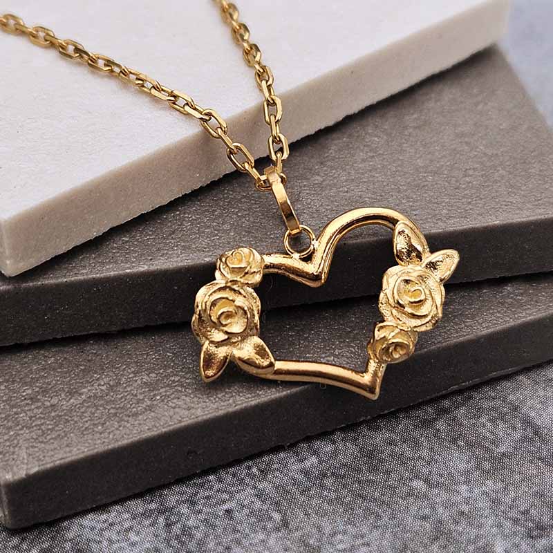 A gold heart-shaped pendant necklace with rose details on the left and right sides, resting on a dark gray textured surface with a white stone block behind it.