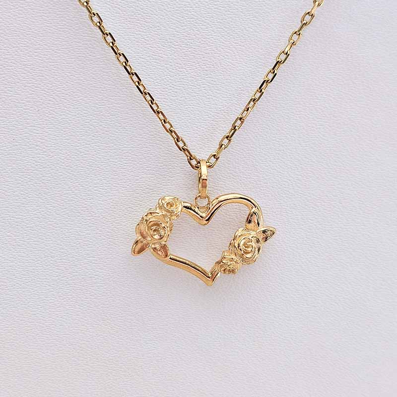 A gold necklace with a heart-shaped pendant adorned with small rose details on both sides, displayed on a textured white background.