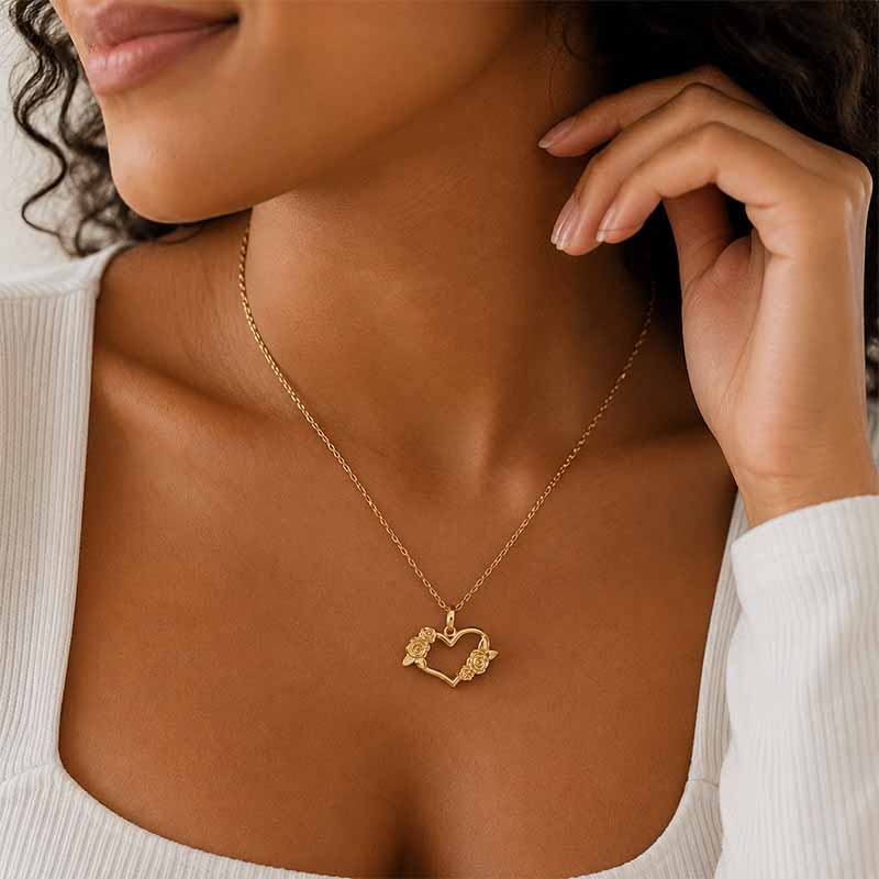 A close-up lifestyle shot of a model wearing a delicate gold chain necklace with a small heart-shaped pendant featuring tiny floral details.