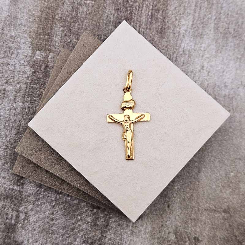 A small gold crucifix pendant displayed on a square cream card, with two gray cards stacked behind it on a textured gray surface.