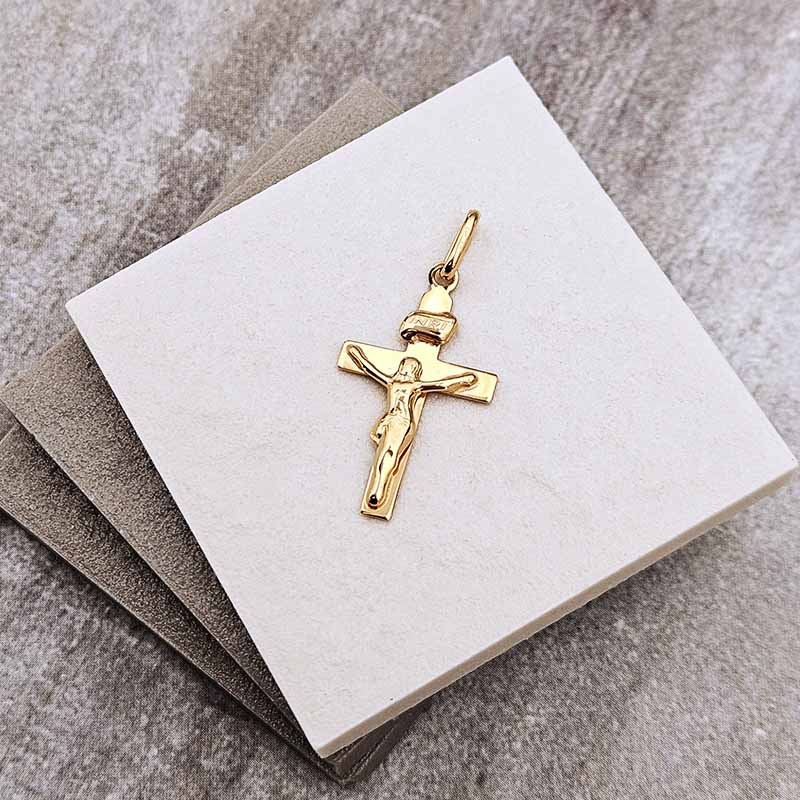 A gold crucifix pendant with a detailed figure of Jesus on a cross, displayed on a white square card over a gray textured surface.