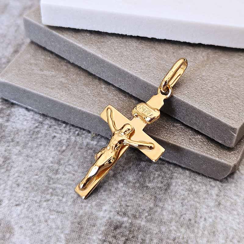 A gold crucifix pendant with a detailed figure of Jesus on a textured gray stone block, with a white block behind it on a gray fabric surface.
