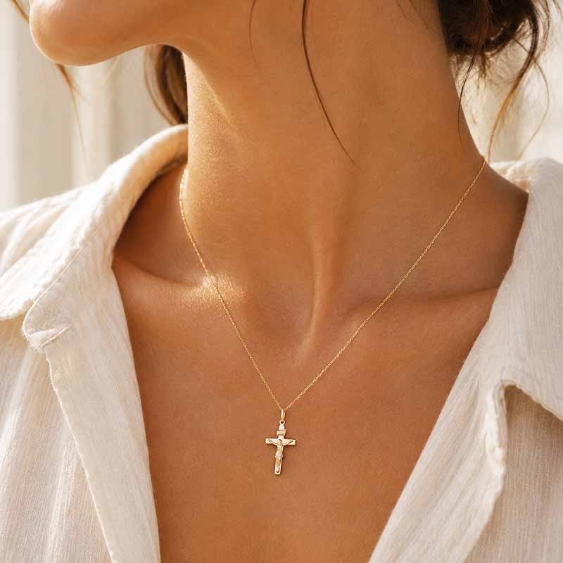 A delicate gold cross pendant necklace worn by a model, shown in a close-up shot of the neck and upper chest wearing a white collared shirt.