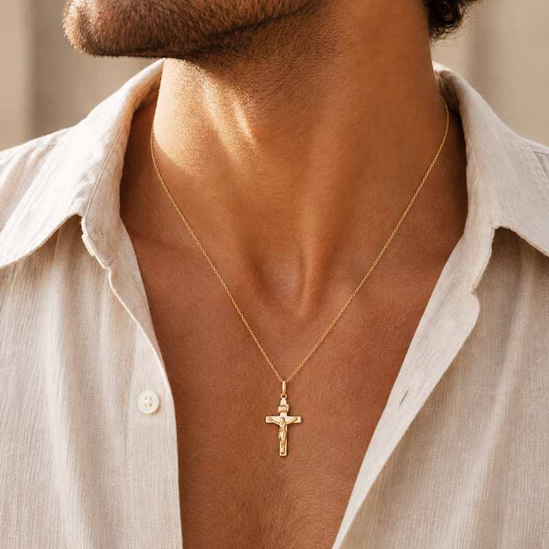 A close-up of a man's neck and chest wearing a delicate gold cross pendant necklace on a fine chain, paired with an open off-white linen shirt.