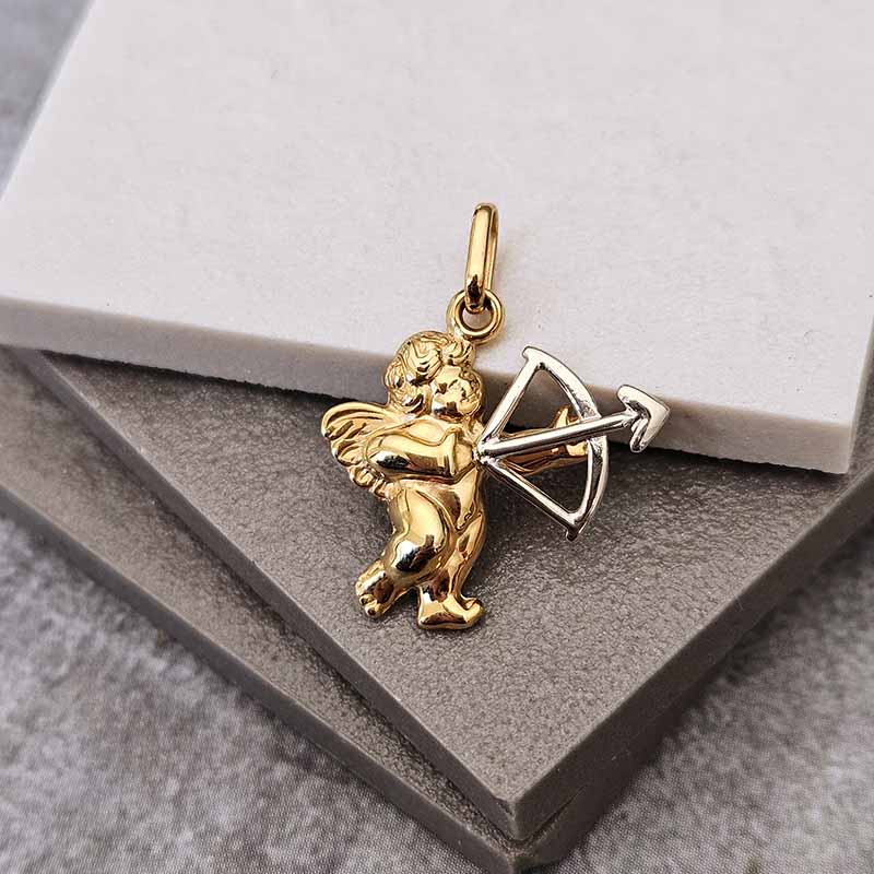 A gold cupid pendant holding a silver bow and arrow, displayed on a gray textured surface with a white marble block.