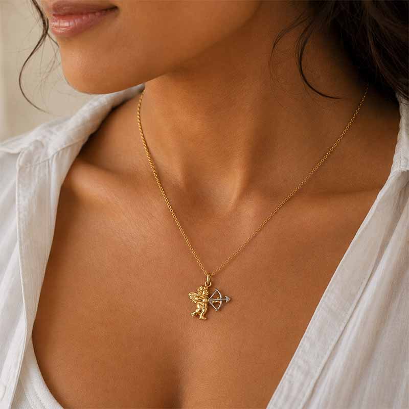 A close-up lifestyle shot of a model wearing a delicate gold chain necklace with a small angel pendant holding a silver bow. The model is wearing a white button-down shirt, and the background is a soft, warm beige.