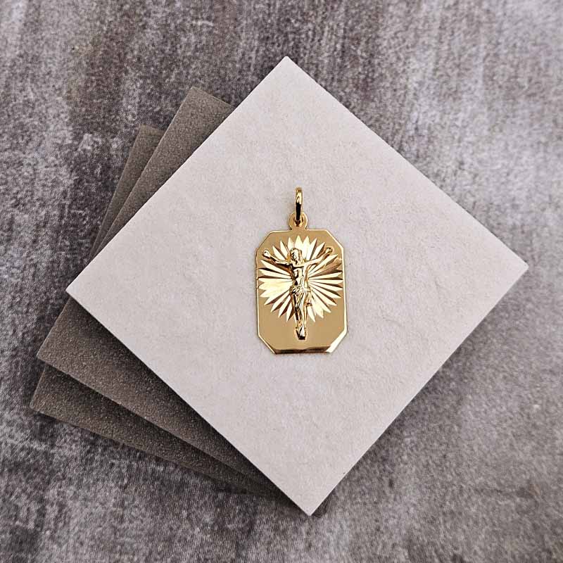 A gold-toned crucifix pendant displayed on a light gray square card, with two darker gray cards stacked behind it on a textured gray surface.