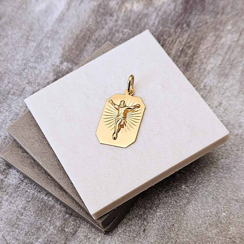 A gold crucifix pendant displayed on a stack of neutral-colored square cards against a textured gray background.