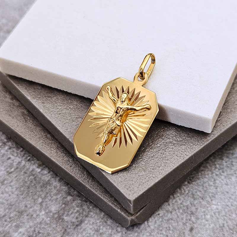 A gold-toned rectangular pendant with a raised figure of Jesus on a sunburst background, displayed on stacked gray and white boxes.
