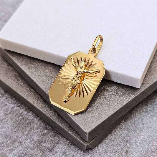 A gold-toned rectangular pendant with a raised figure of Jesus on a sunburst background, displayed on stacked gray and white boxes.