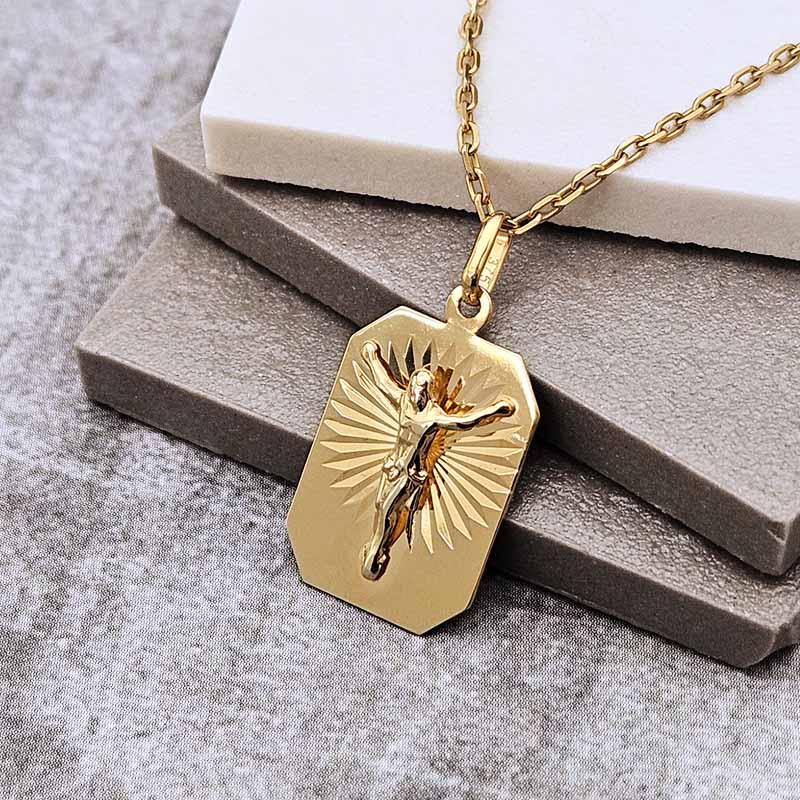 A gold crucifix pendant necklace on a matching chain, displayed on stacked gray and white blocks over a textured gray surface.