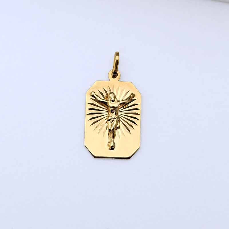 A gold crucifix pendant charm with a raised figure of Jesus on a rectangular tag-shaped background, shown on a plain light background.