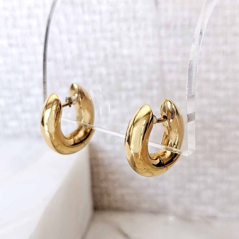 A pair of polished gold huggie hoop earrings displayed on a clear acrylic stand against a soft, textured light gray background.