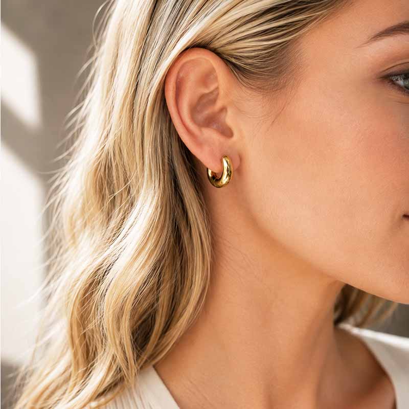 A close-up of a woman's ear wearing a small gold hoop earring, with her blonde hair and skin visible against a softly blurred neutral background.