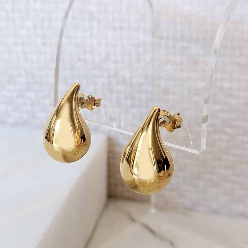 A pair of gold teardrop-shaped stud earrings displayed on a clear acrylic stand against a light textured background.