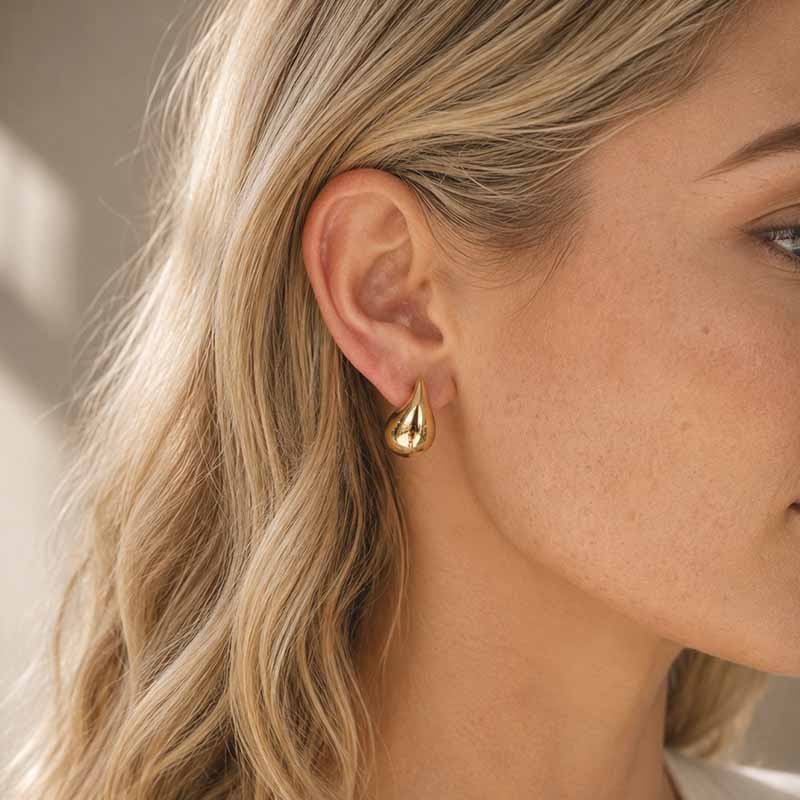 A close-up of a woman's ear wearing a small gold teardrop-shaped stud earring, with blonde hair and soft natural light in the background.