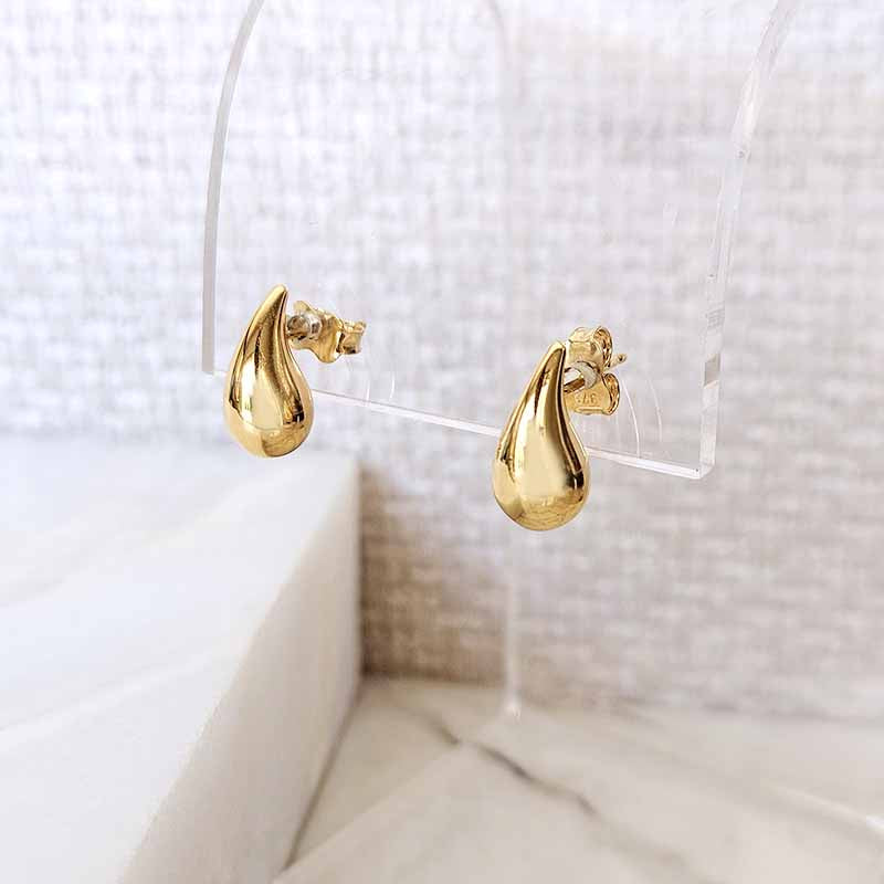 A pair of gold teardrop-shaped stud earrings displayed on a clear acrylic stand against a soft, textured light gray background.