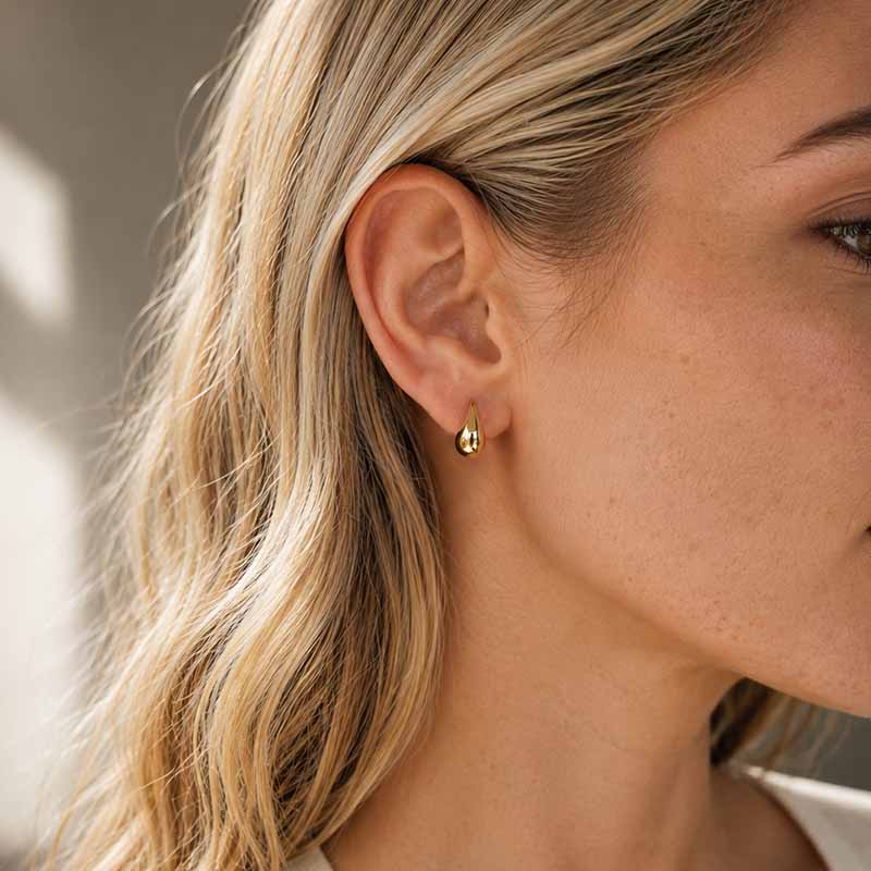 A close-up of a woman's ear wearing a small gold teardrop stud earring, with soft natural light and a blurred neutral background.