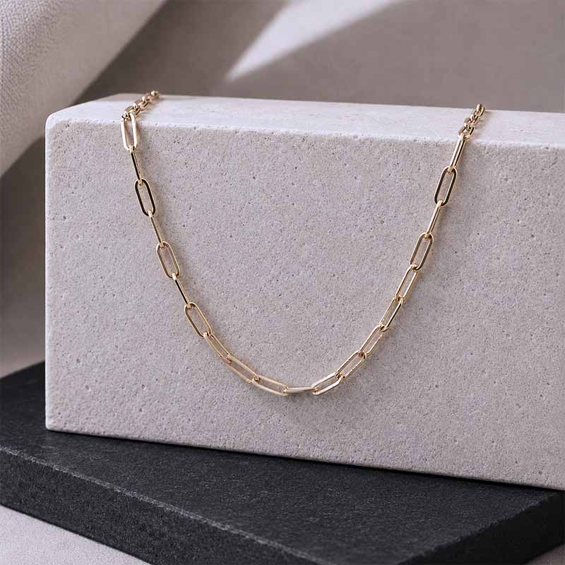 A gold paperclip chain necklace draped over a textured light gray rectangular display block, with a dark gray surface underneath.