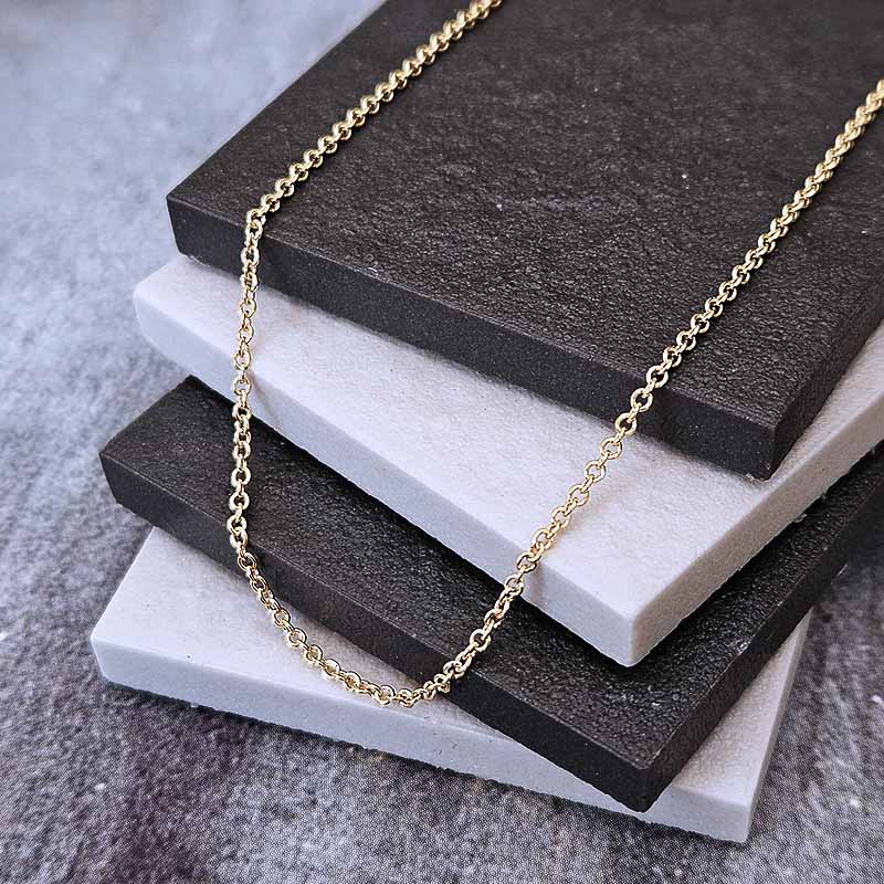 A delicate gold chain necklace draped over a stack of three rectangular display blocks in dark charcoal, light gray, and white, set against a textured gray background.