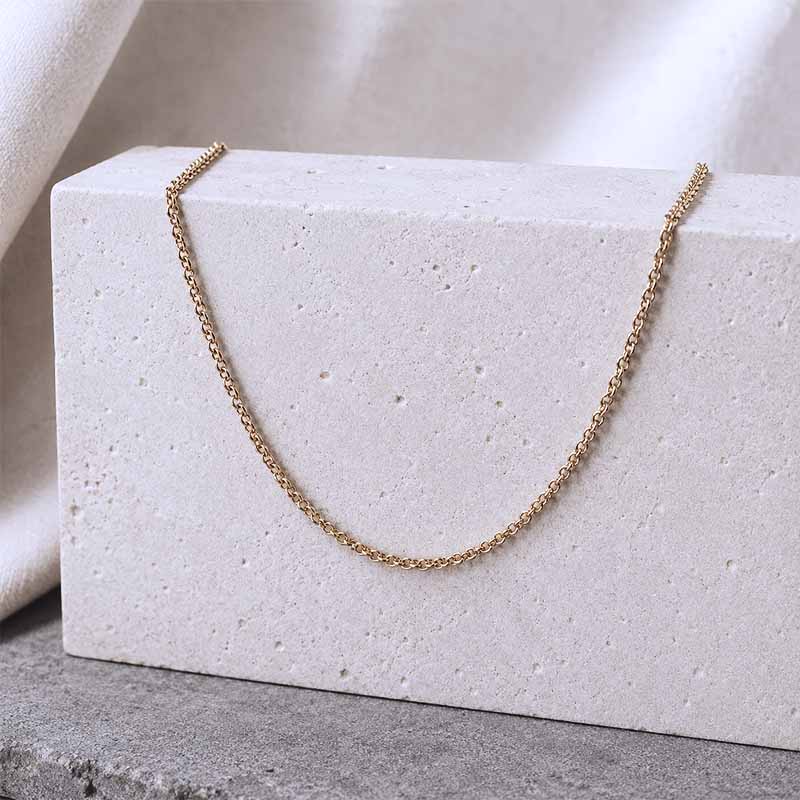 A delicate gold chain necklace draped over a textured white stone block, set against a soft beige fabric backdrop and a gray surface.