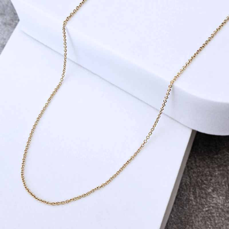 A delicate gold chain necklace draped over a white display block, showing its fine link structure against a clean white background with a dark textured surface in the corner.