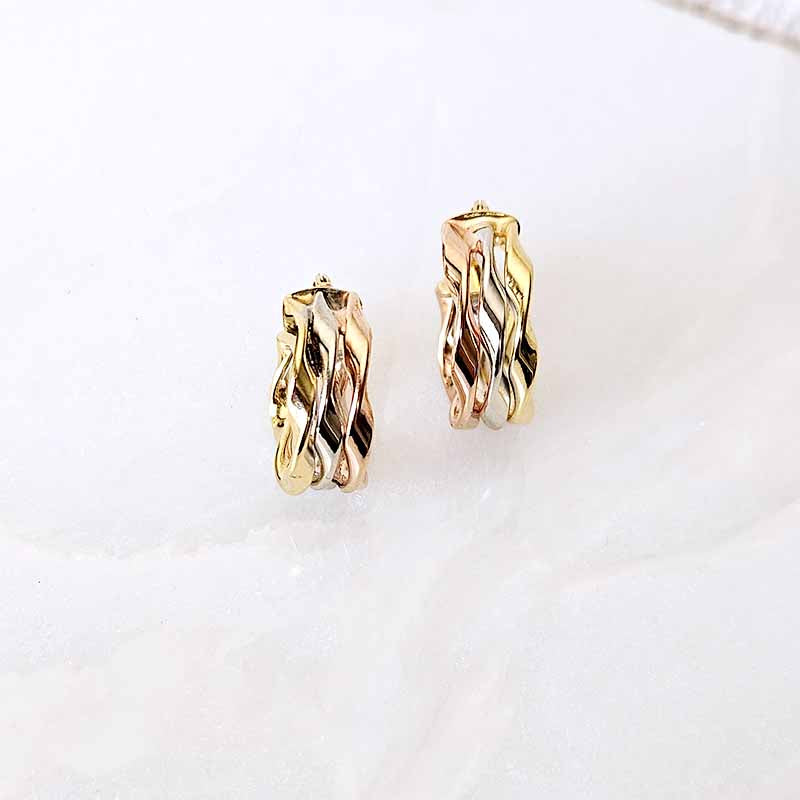 A pair of twisted huggie hoop earrings in a two-tone gold and rose gold finish, displayed on a soft, light-colored marble surface.