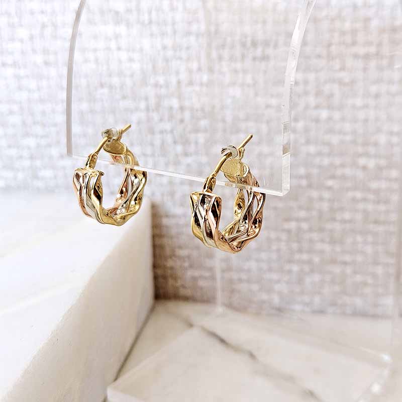 A pair of gold hoop earrings with a twisted, textured design, displayed on a clear acrylic stand against a neutral textured background.