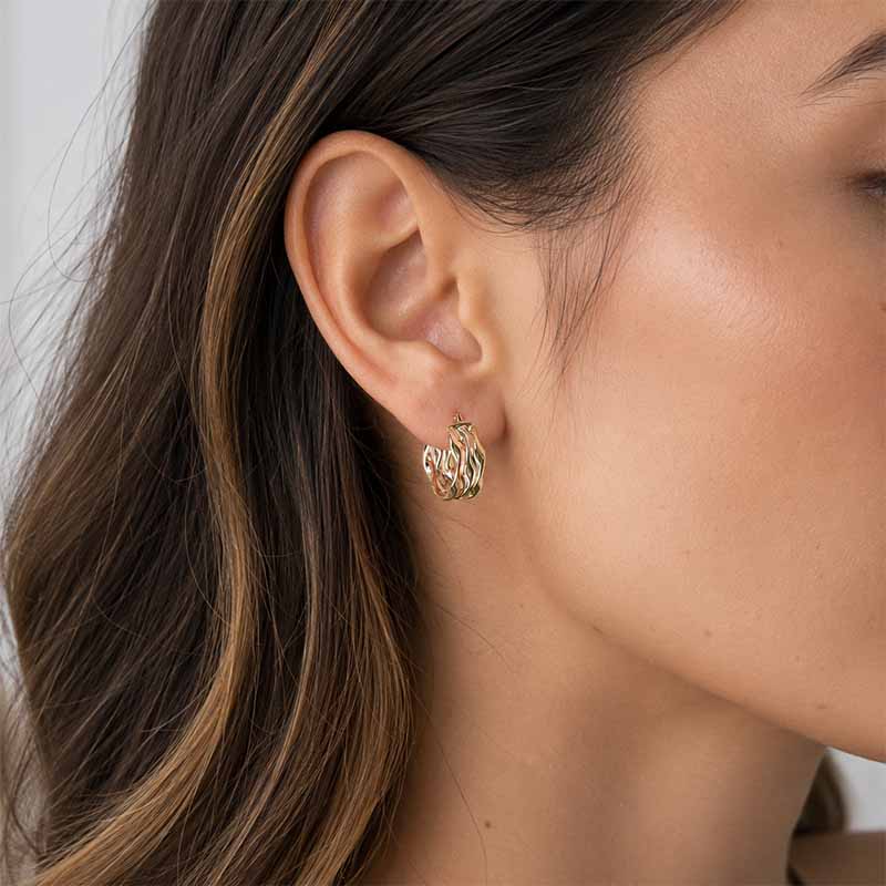A close-up of a woman's ear wearing a small gold huggie hoop earring with a textured, twisted design.