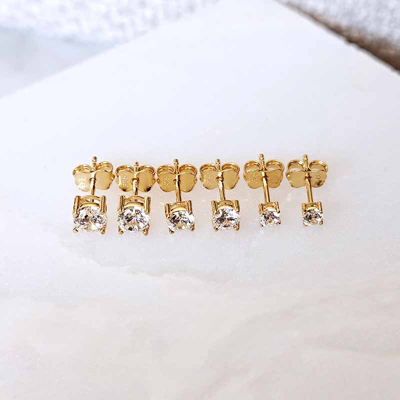 Six pairs of gold stud earrings with clear round-cut gemstones, arranged in a row on a white textured surface.