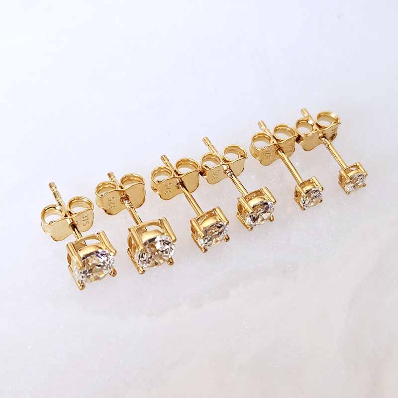 Six pairs of gold stud earrings with clear gemstones, arranged in a row on a plain white background.