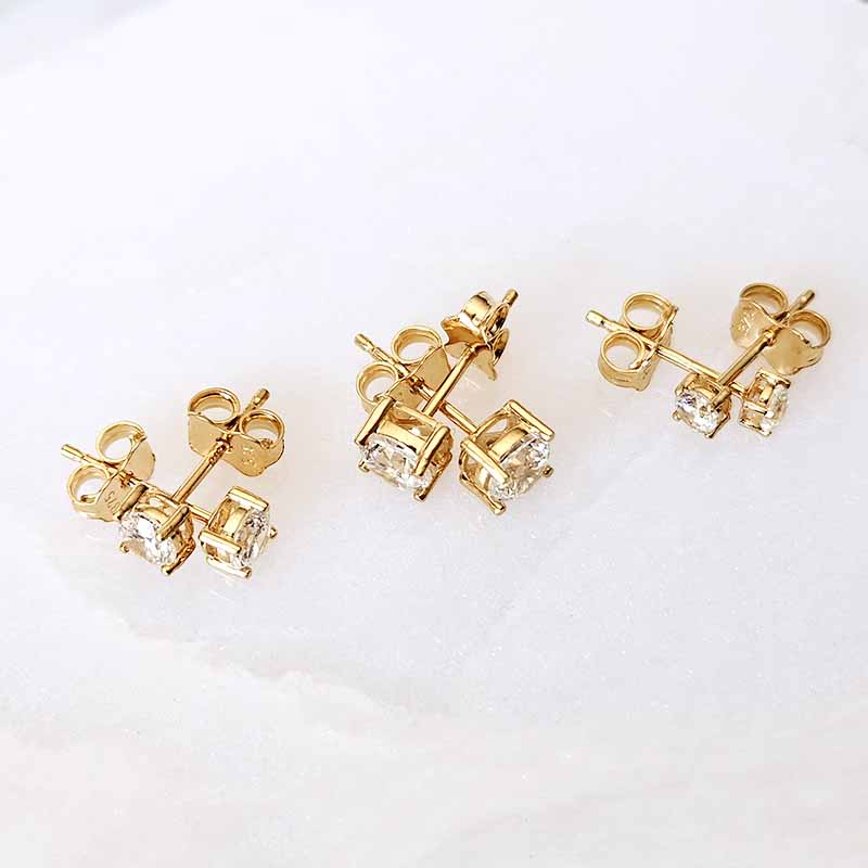 Three pairs of gold stud earrings with clear gemstones, displayed on a white textured surface.