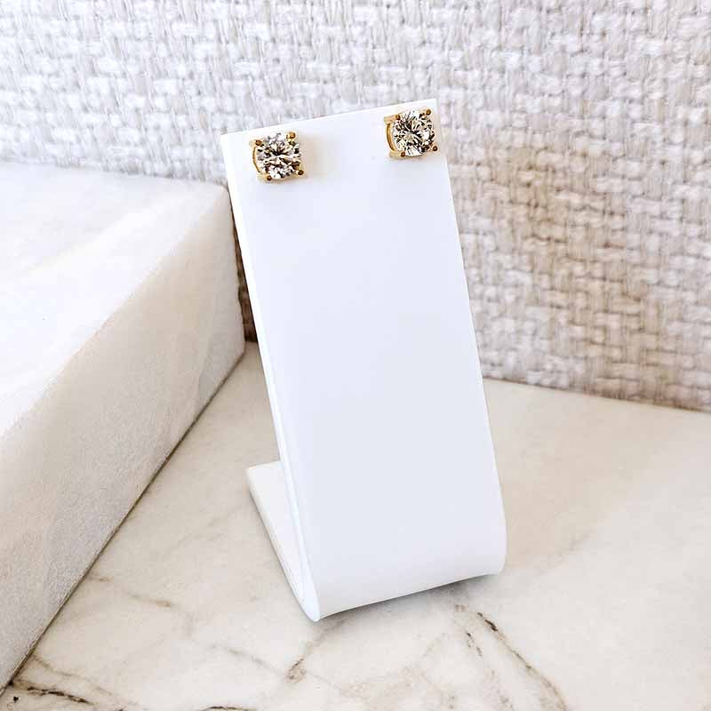 A pair of gold stud earrings with clear stones, displayed on a white stand on a marble surface with a textured woven wall background.