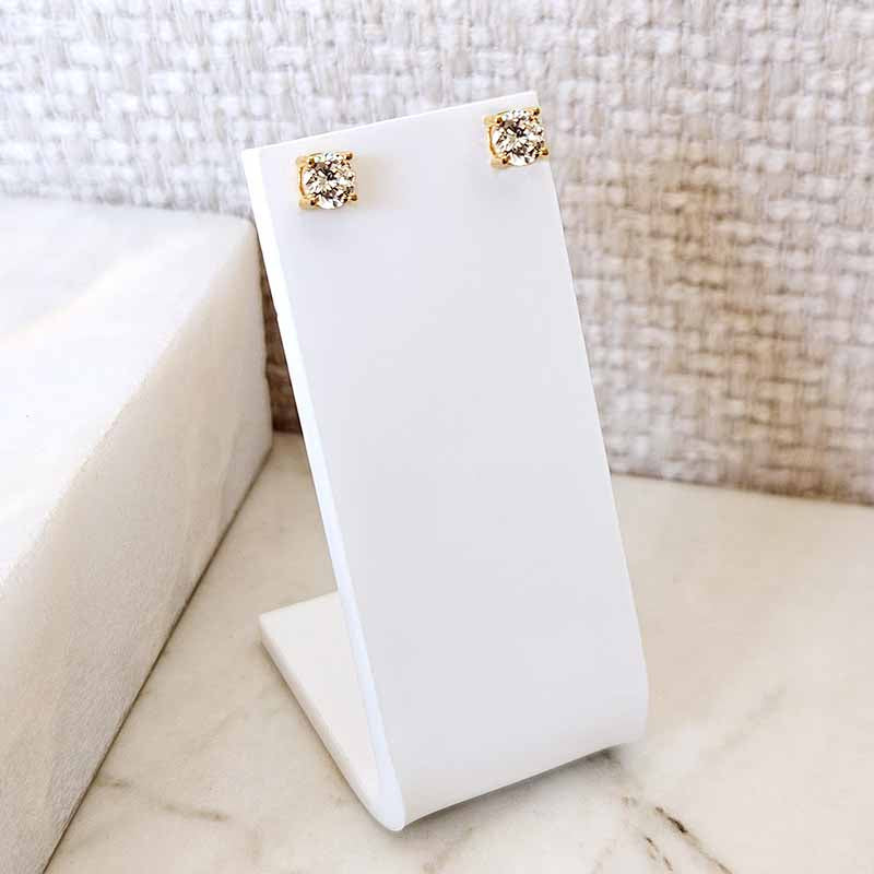 A pair of gold stud earrings with clear gemstones displayed on a white jewelry stand, set against a neutral textured background.
