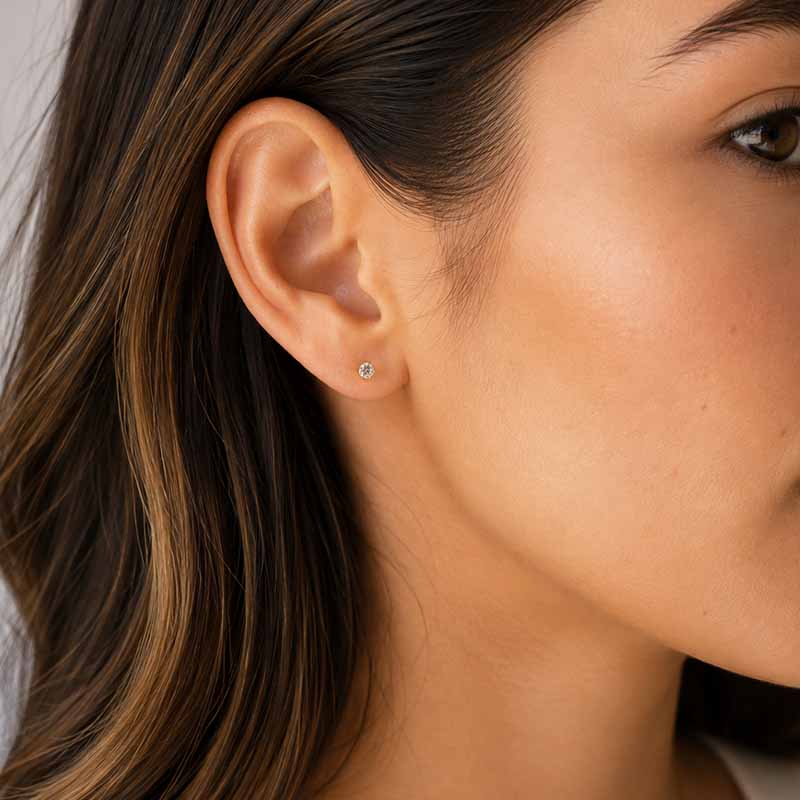 A close-up of a woman's ear wearing a tiny round stud earring with a clear gemstone in a silver setting.