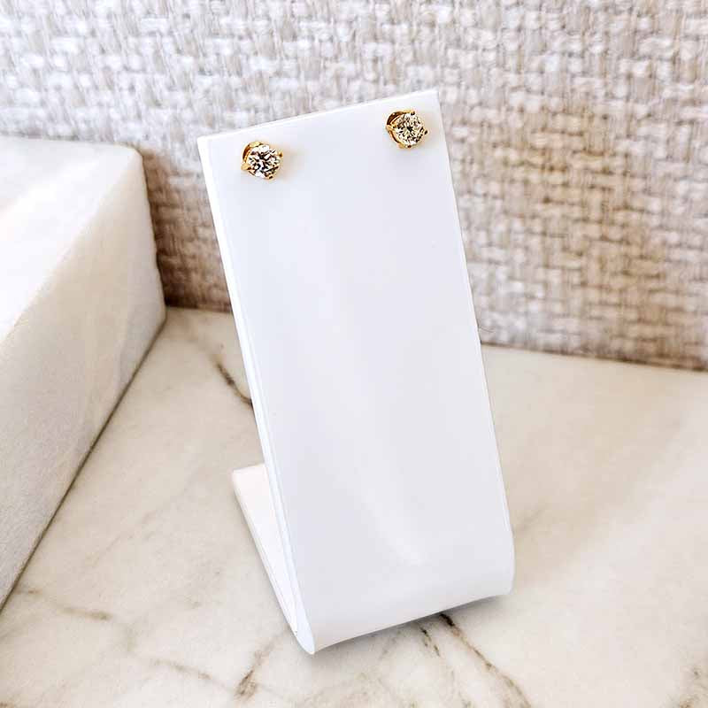 A pair of small gold stud earrings displayed on a white stand, set on a light marble surface against a woven beige background.