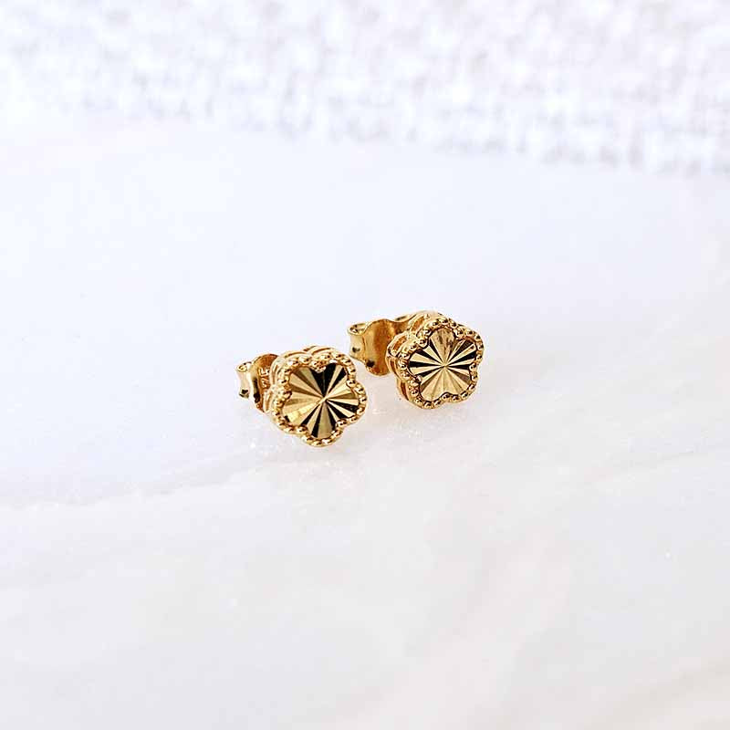 A pair of gold stud earrings with a textured, starburst-like floral face and a scalloped edge, resting on a white marble surface.