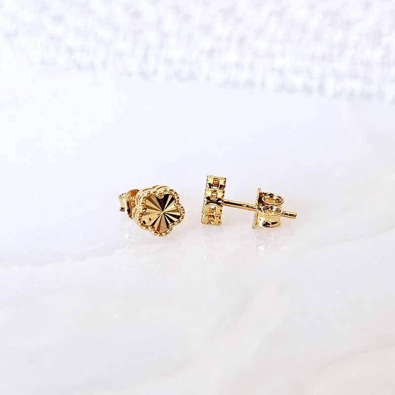 A pair of gold stud earrings, one shown from the front with a textured, flower-like faceted design and the other from the side/back showing the post and butterfly back, resting on a white marble surface.
