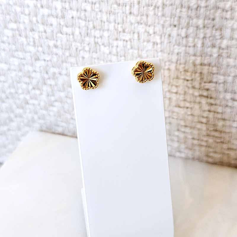 A pair of small gold-tone stud earrings with a textured, flower-like starburst design, displayed on a white card stand against a neutral textured background.