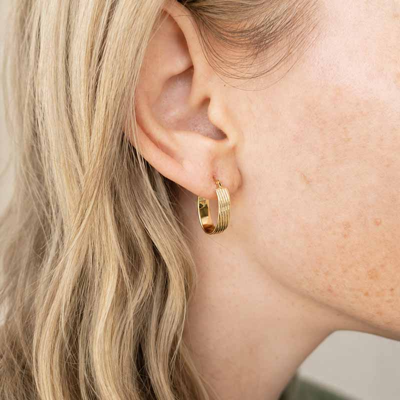A close-up of a model's ear wearing a small gold hoop earring with a textured, ribbed outer band.