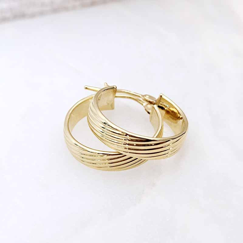 A pair of gold hoop earrings with a textured, ridged outer band and a smooth inner band, displayed on a light marble surface.