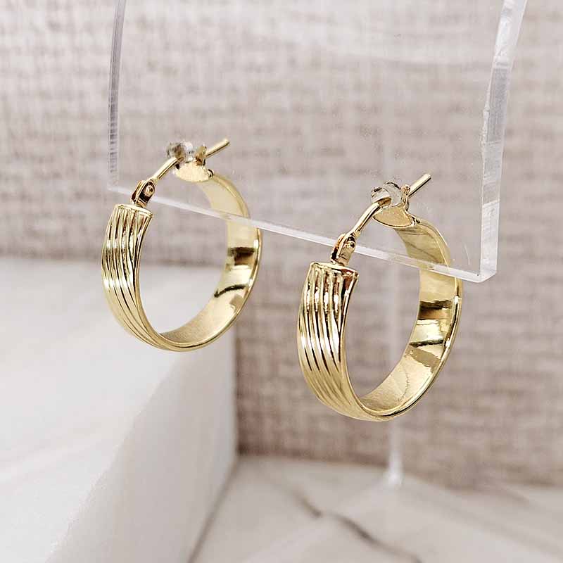 A pair of gold hoop earrings with a textured, ridged inner band and a smooth outer edge, displayed on a clear acrylic stand against a neutral beige background.
