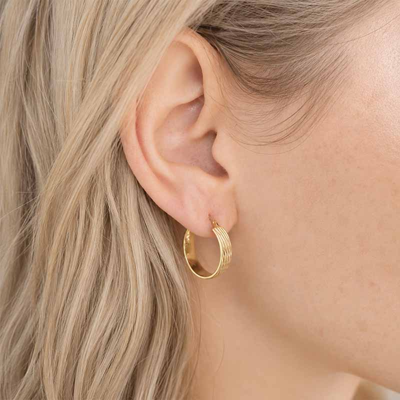 A close-up of a model's ear wearing a small gold hoop earring with a textured, wavy band design.