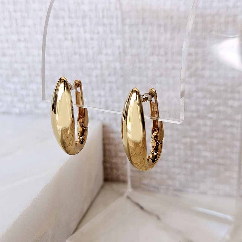 A pair of polished gold huggie hoop earrings displayed on a clear acrylic stand against a soft, textured light gray background.