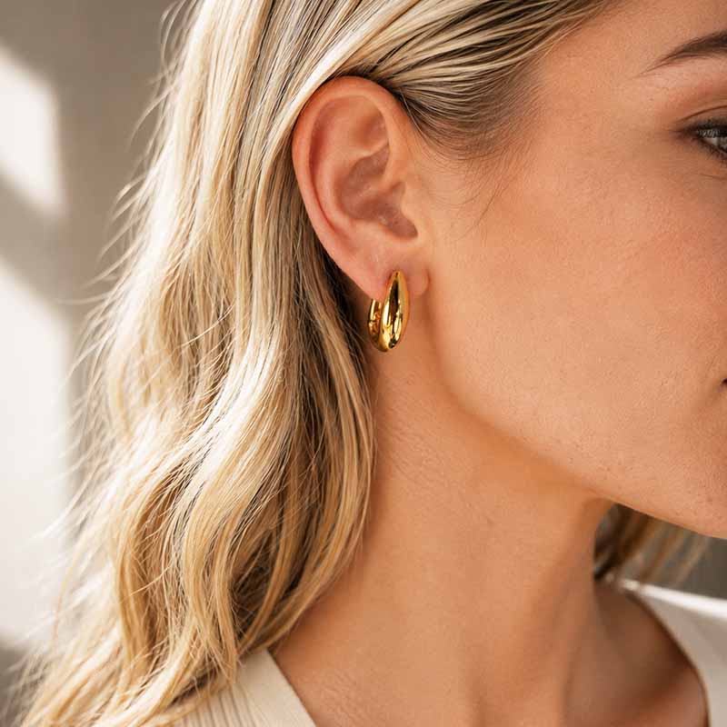 A close-up of a woman's ear wearing a small polished gold hoop earring, with blonde hair and a neutral indoor background.