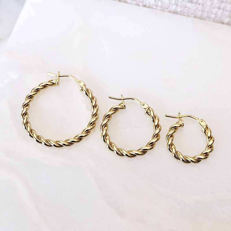 Three gold-toned twisted hoop earrings in different sizes arranged on a light off-white surface.