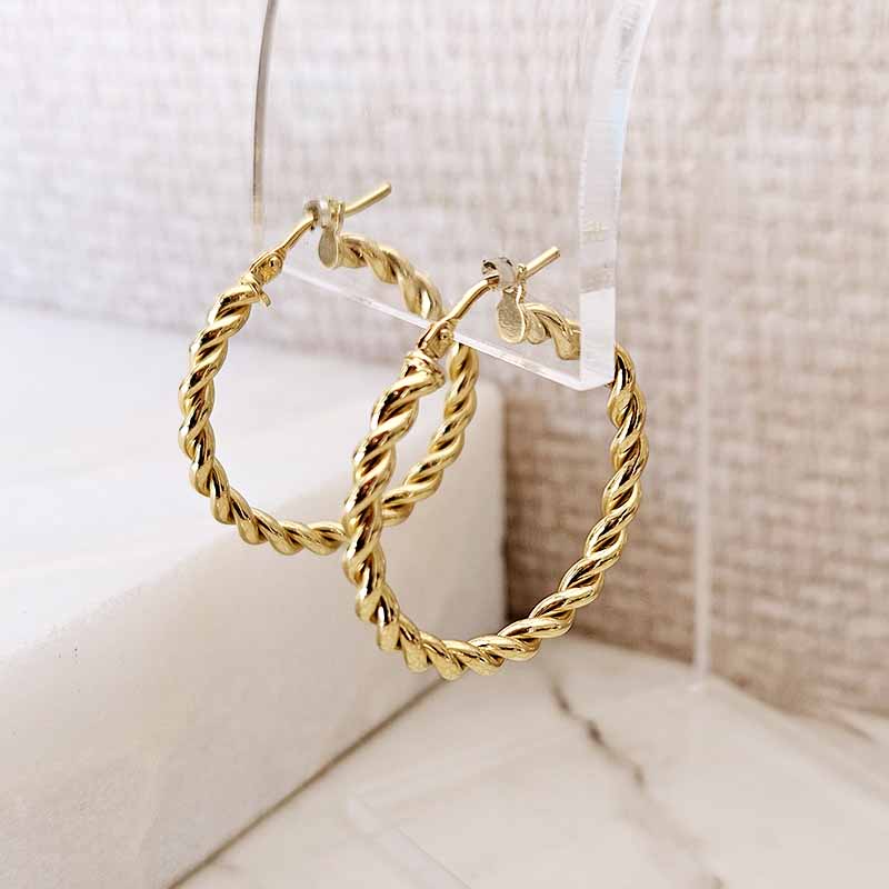 A pair of gold hoop earrings with a twisted rope texture, hanging from a clear acrylic display stand against a neutral beige textured background.